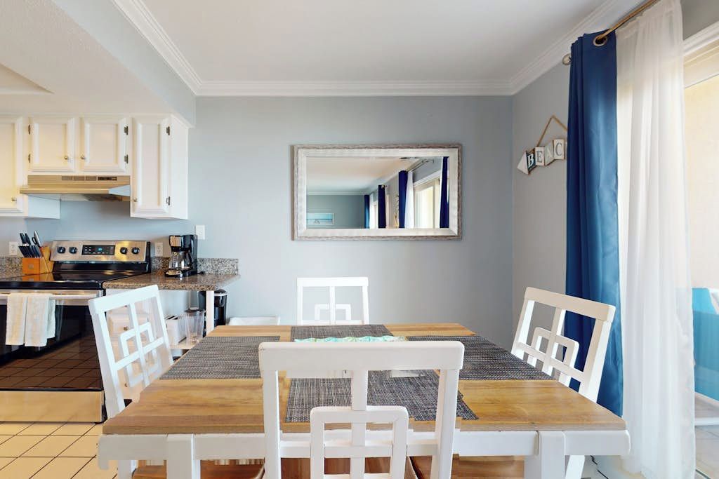 A dining area with a wooden table and white chairs, a kitchen with white cabinets, and a mirror on the wall.