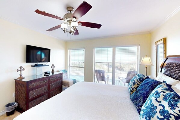 A hotel room with a bed, a ceiling fan, a television, a dresser, and a balcony with chairs. - Suntide II Condominiums #106 (South Padre Island)