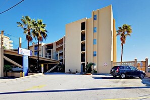 A multi-story building with a parking area in front, palm trees, and a sign for a business. - Suntide II Condominiums #106 (South Padre Island)