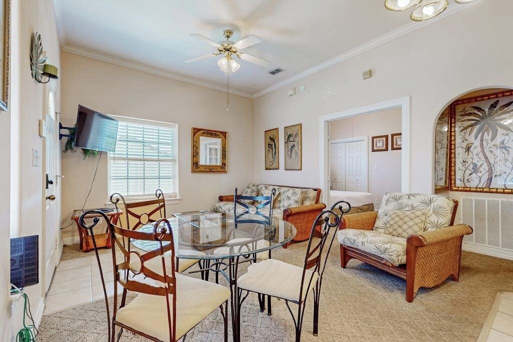 Sala de estar com mesa de jantar de vidro, cadeiras de vime, ventilador de teto e obras de arte nas paredes.