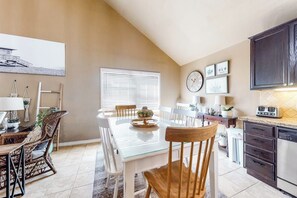 A dining area with a glass table, white chairs, and a wooden chair. There is a window with blinds, a clock on the wall, and a kitchen area with cabinets and appliances. - Aries Chateau - Private Pool, Half Block to Beach (South Padre Island)