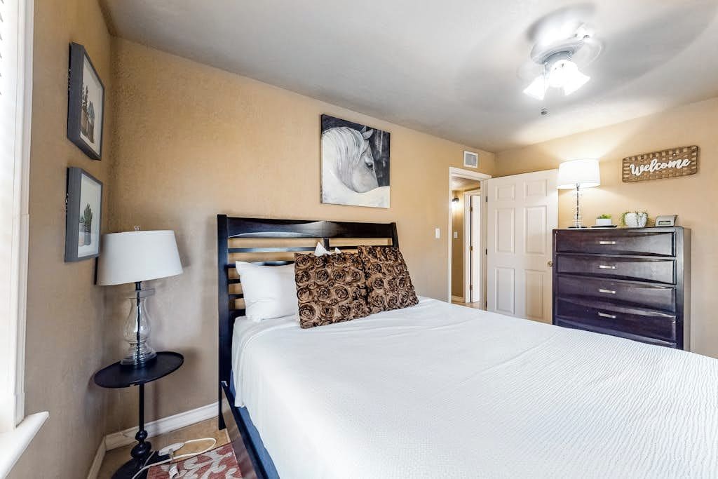 A hotel room with a bed, bedside lamp, a wall-mounted painting, a dresser, and a door leading to another room.