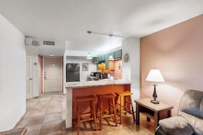 A compact kitchen with a central island, bar stools, a small table with a lamp, and a sofa in a living area. - Gulfview I #207 - Bay Views, Pool, Walk to Beach (South Padre Island)