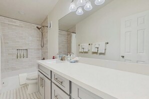 Un baño con lavamanos blanco, espejo y ducha. Hay toallas colgadas en la pared y un inodoro.