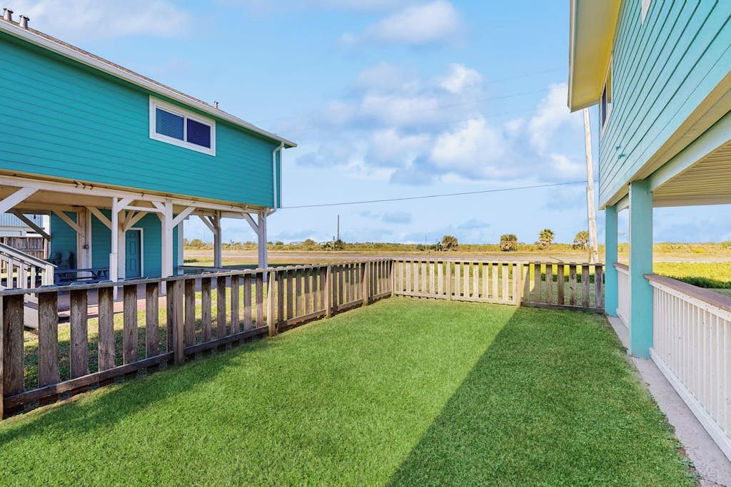 A fenced backyard with a grassy lawn, a wooden deck, and a house with a blue exterior.