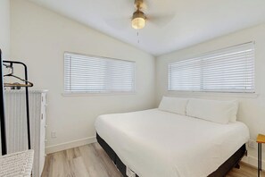 Ein Schlafzimmer mit einem Bett, zwei Fenstern mit Jalousien, einem Deckenventilator und einem kleinen Tisch.