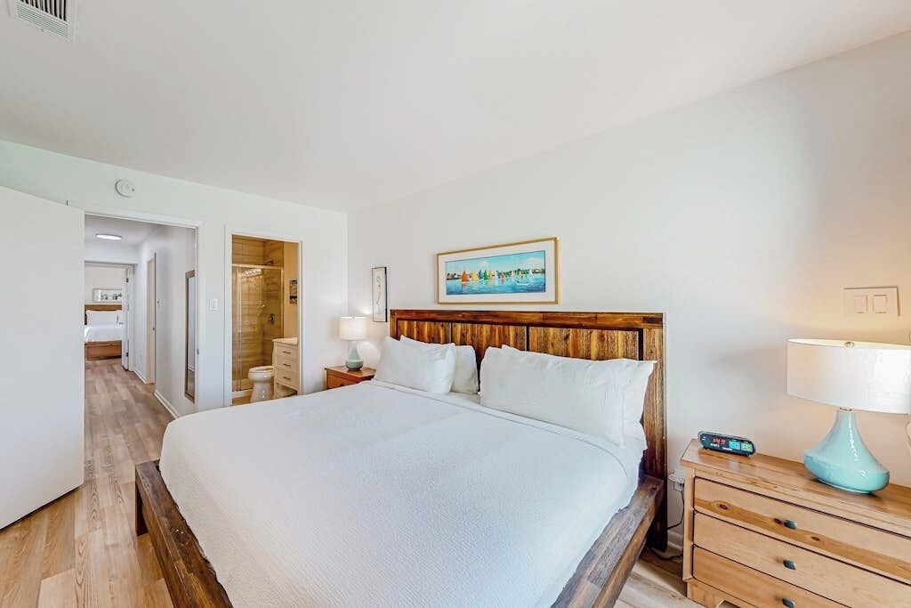 Een hotelkamer met een groot bed, nachtkastjes, een houten hoofdbord, een schilderij aan de muur en een badkamer zichtbaar door een geopende deur.