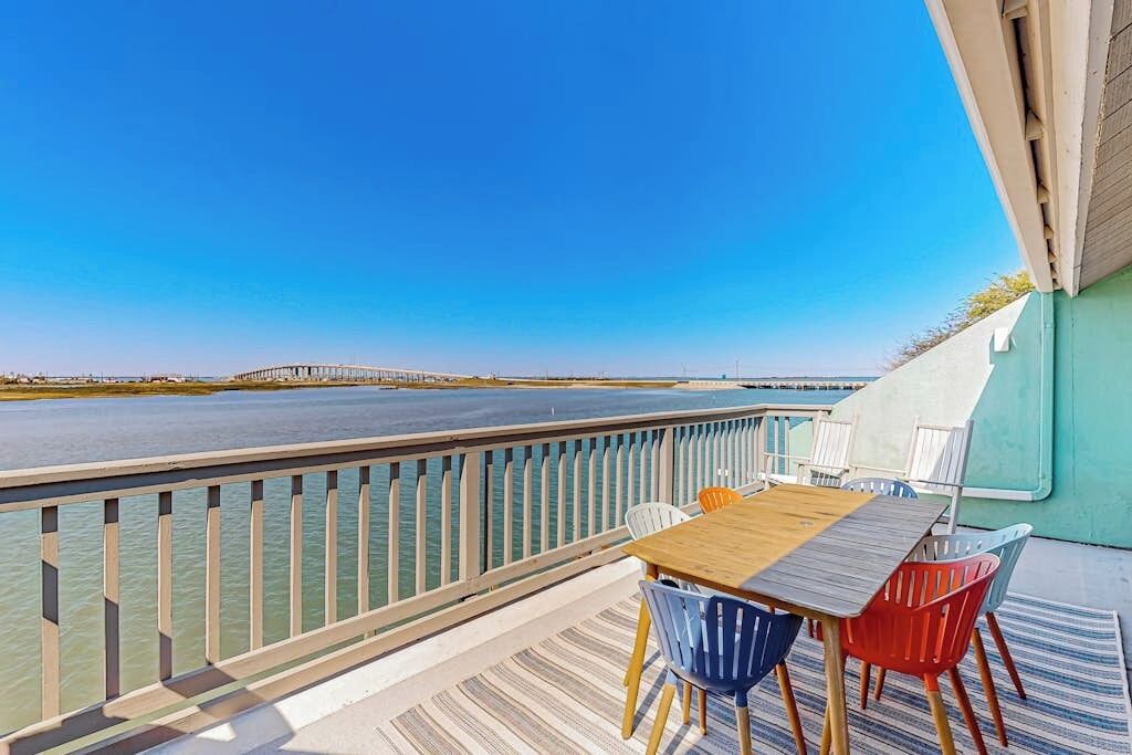 A deck with a table and chairs overlooking a body of water and a distant bridge.