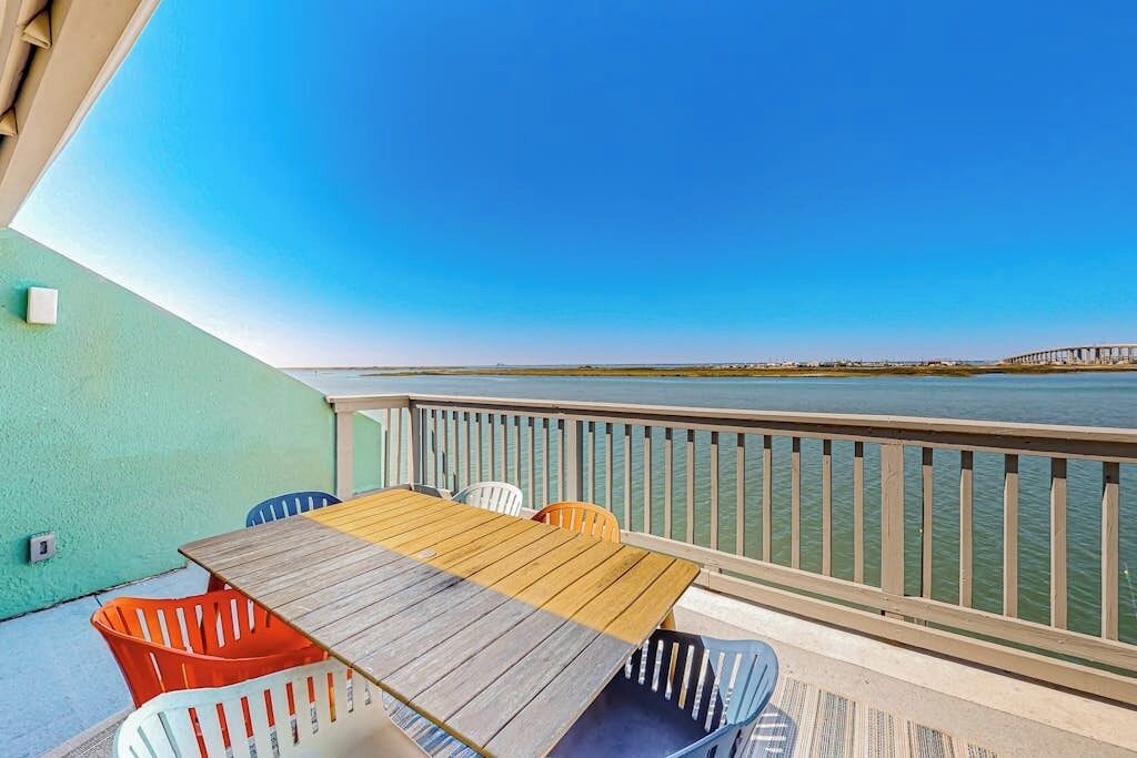 Une terrasse en bois avec une table et des chaises, donnant sur une étendue d’eau et un pont situé au loin.