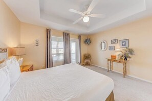A hotel room with a large bed, a ceiling fan, a window with curtains, a small table with a lamp, and a wall-mounted mirror. - Cabana Street Unit B (Corpus Christi)