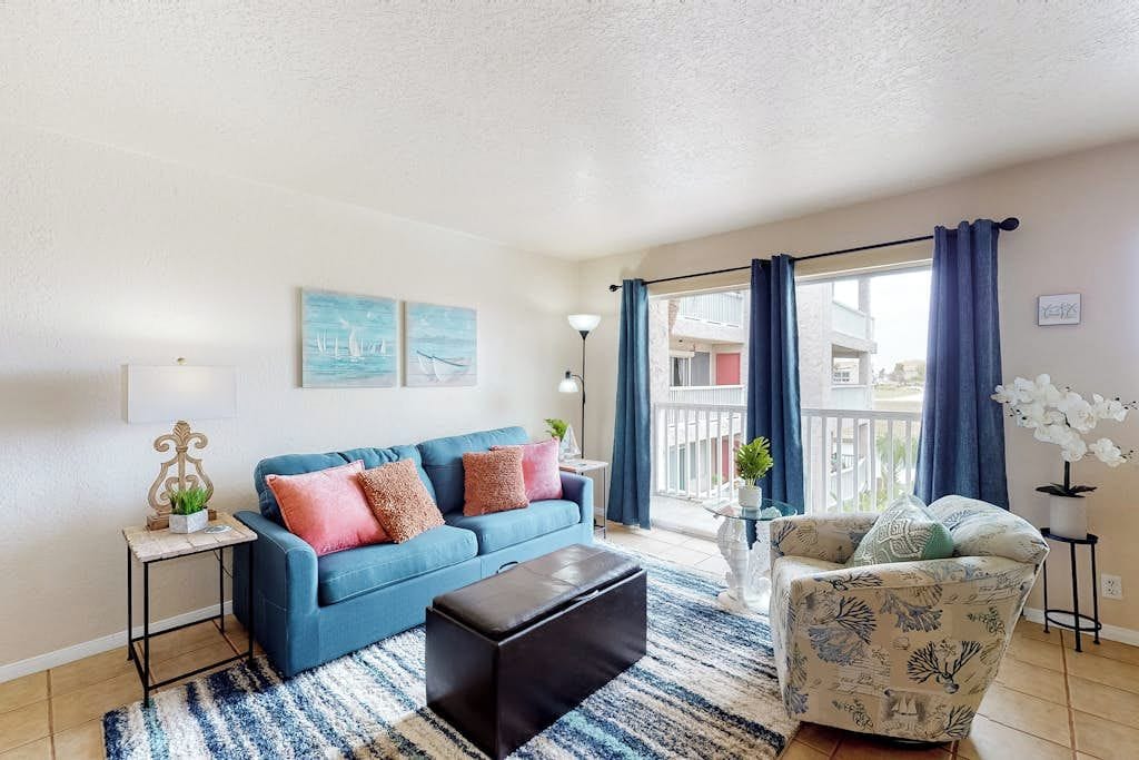 Una sala de estar con un sofá azul, un sillón estampado, un reposapiés negro y vistas al balcón exterior.