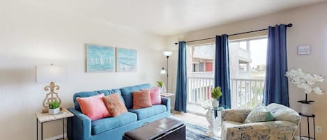 Una sala de estar con un sofá azul, un sillón estampado, un reposapiés negro y vistas al balcón exterior.