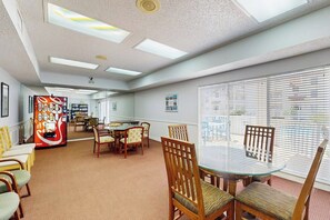 A spacious room with a round glass table, wooden chairs, a vending machine, and large windows with blinds. - Beach Club 326 - Pool, 2 Blocks to Beach, Dogs OK (Corpus Christi)