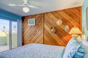 Camera da letto con parete in legno a spina di pesce, ventilatore a soffitto, letto con biancheria a motivi decorativi e vista attraverso una porta scorrevole in vetro.