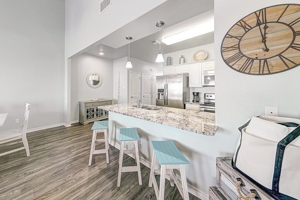 Una cocina moderna con una isla central, taburetes de bar, iluminación colgante y un reloj de pared grande.