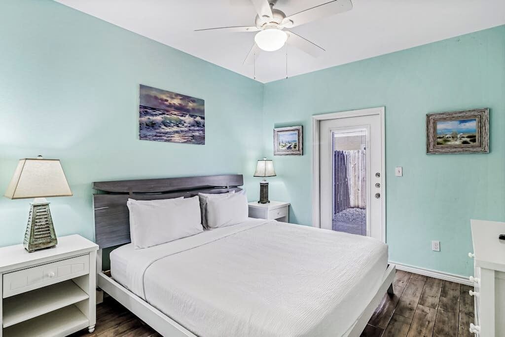 A bedroom with a bed, a nightstand, a lamp, a ceiling fan, and a door leading to another room.