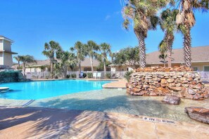 A clear swimming pool with a 9-inch depth marker, surrounded by palm trees and a stone wall. - Palm Bay 407 - Heated Pool & Covered Patio (Corpus Christi)