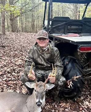 A person in camouflage attire holding antlers, sitting next to a vehicle in a wooded area.