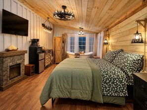 A cozy room with a fireplace, a bed with patterned bedding, a wooden ceiling, and a television mounted on the wall.