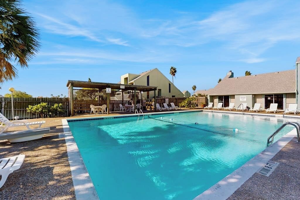 Une piscine dotée de chaises longues et d’un espace de repos couvert, entourée d’immeubles et de palmiers.
