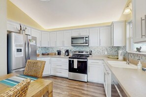 A modern kitchen with white cabinets, stainless steel appliances, and a dining area with a wicker chair. - The Lazy Bay - Canal-Front Home with Boat Dock (Rockport)