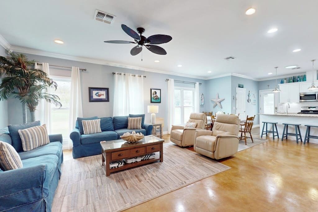 A spacious living room with a ceiling fan, a dining area with a kitchen in the background, and a large potted plant.