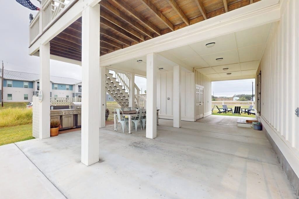 A covered outdoor area with a concrete floor, a staircase leading to an upper level, and a view of a residential area in the background.