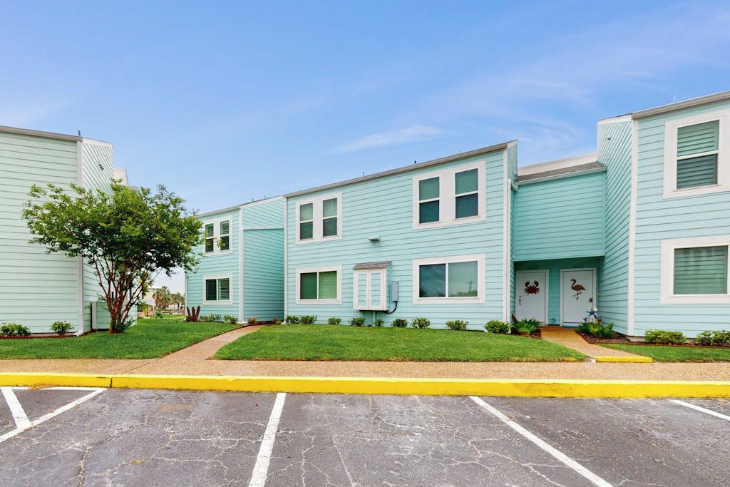 A row of light blue townhouses with a grassy front yard and a parking lot in front.