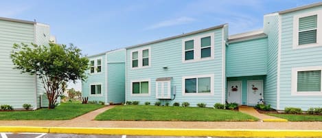A row of light blue townhouses with a grassy front yard and a parking lot in front.