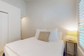 A bedroom with a bed, pillows, a bedside table with a lamp, and a window with blinds. - Pelican Point - Waterfront, Pool & Dock Access (Rockport)