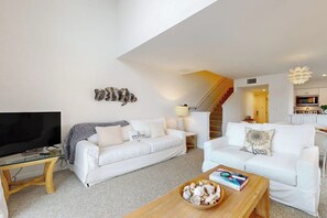 A modern living room with a flat-screen TV, two white sofas, a coffee table with books and a bowl, and a staircase leading to a kitchen. - Pelican Point - Waterfront, Pool & Dock Access (Rockport)