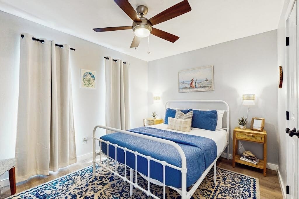 Quarto com cama, cortinas, ventilador de teto, mesa de cabeceira com abajur e pintura na parede.