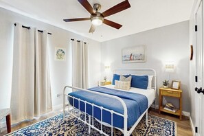 A bedroom with a bed, curtains, a ceiling fan, a bedside table with a lamp, and a wall painting. - The Bluff Breeze - Canal-Front with Large Yard (Corpus Christi)