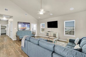 A living room with a sectional sofa, a flat-screen TV, a ceiling fan, and a window with blinds. - Blue Pelican - Bayfront, Fenced Yard & Balconies (Corpus Christi)