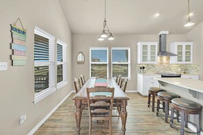A dining room with a long table and chairs, a kitchen area with white cabinets and a brick backsplash, and a hanging light fixture. - Blue Pelican - Bayfront, Fenced Yard & Balconies (Corpus Christi)