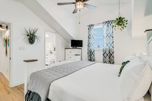 Ein Schlafzimmer mit einem Bett, einem Deckenventilator, einem Fernseher, einem Schminktisch, einem Spiegel und einem Fenster mit Vorhängen.