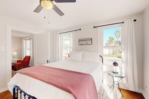 A bedroom with a bed, a ceiling fan, a window with curtains, a small table with a lamp, and a chair. - Kay's Kottage - Fenced Yard, Near Beach & Seawall (Galveston)