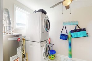 A laundry room with a stacked washer and dryer, a water heater, and a wall-mounted drying rack with bags. - Kay's Kottage - Fenced Yard, Near Beach & Seawall (Galveston)