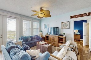 Ein Wohnzimmer mit einer Couch, einem Couchtisch und einem Fernseher. Es gibt einen Deckenventilator, ein Schild mit der Aufschrift 'BEACH HOUSE' und eine Tür, die nach draußen führt.