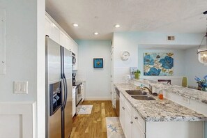 A kitchen with white cabinets, a stainless steel refrigerator, a granite countertop, a sink, and a clock on the wall. - Pointe West Resort - Seas the Day (Galveston)
