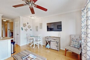 Ein Wohnzimmer mit Deckenventilator, einem an der Wand montierten Fernseher, einem Essbereich mit weißen Stühlen und einem kleinen Tisch.