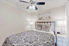 Een slaapkamer met een bed, een plafondventilator, een nachtkastje, een lamp en een schilderij aan de muur.