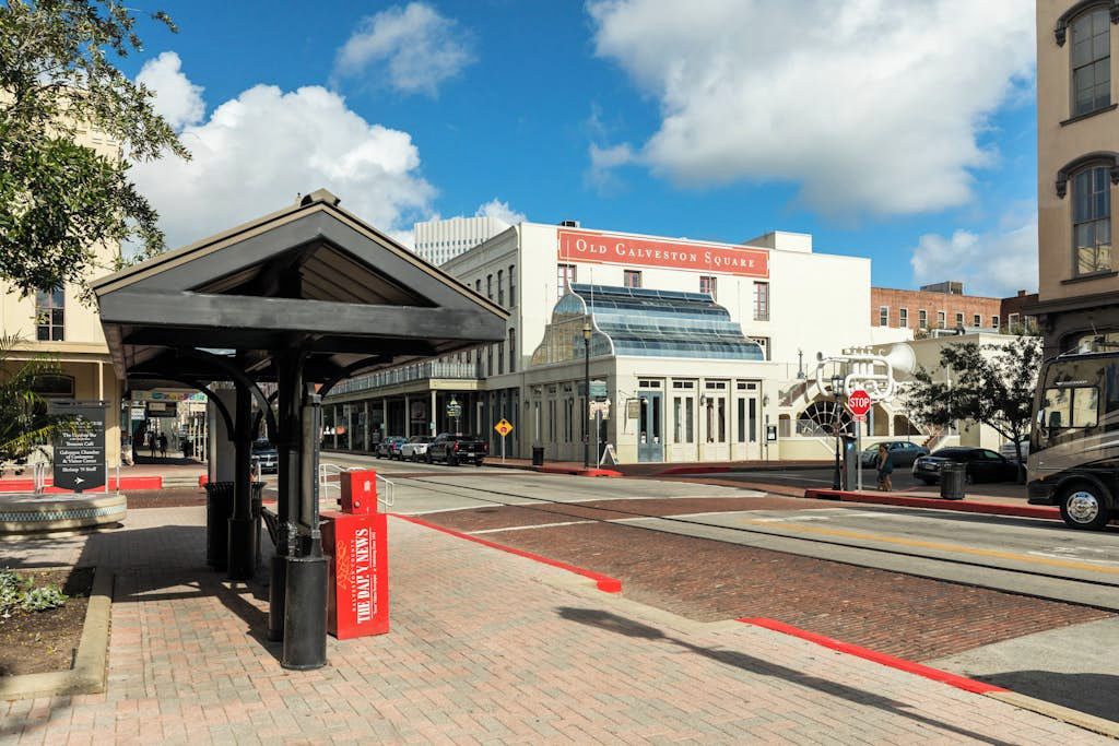 A street view with a covered bus stop, a building labeled 'Old Galveston Square,' and a clear blue sky.