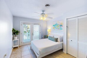 A bedroom with a bed, a ceiling fan, a window with blinds, a door, and a wall-mounted artwork. - Phoebe's Beach House (South Padre Island)