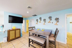 A dining area with a table and chairs, a television mounted on the wall, and a cabinet with a decorative item on top. - Phoebe's Beach House (South Padre Island)