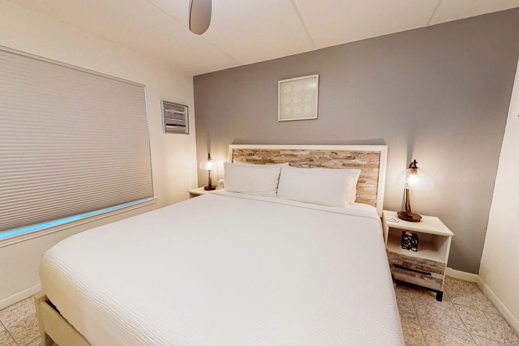 A bedroom with a large bed, two bedside tables with lamps, a headboard with a wood-like design, a window with blinds, and a small picture on the wall.