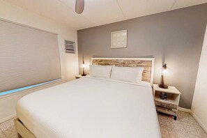 Ein Schlafzimmer mit einem großen Bett, zwei Nachttischen mit Lampen, einer Bettkopfteil mit Holzoptik, einem Fenster mit Jalousien und einem kleinen Bild an der Wand.