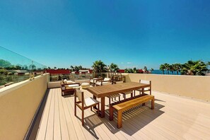 A wooden deck with outdoor furniture, including a table and chairs, overlooking a body of water with palm trees in the background. - Shibui Home - Private Pool, Steps to Sand (South Padre Island)