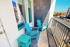 A balcony with two turquoise Adirondack chairs and a small table, overlooking a street with parked cars and buildings. - Lantana Cove #203 - Gulf Views, Across from Beach (South Padre Island)