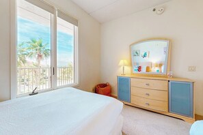 A bedroom with a bed, a dresser, a mirror, and a window with a view of palm trees. - Palm Frond Sanctuary, Unit 302 (South Padre Island)